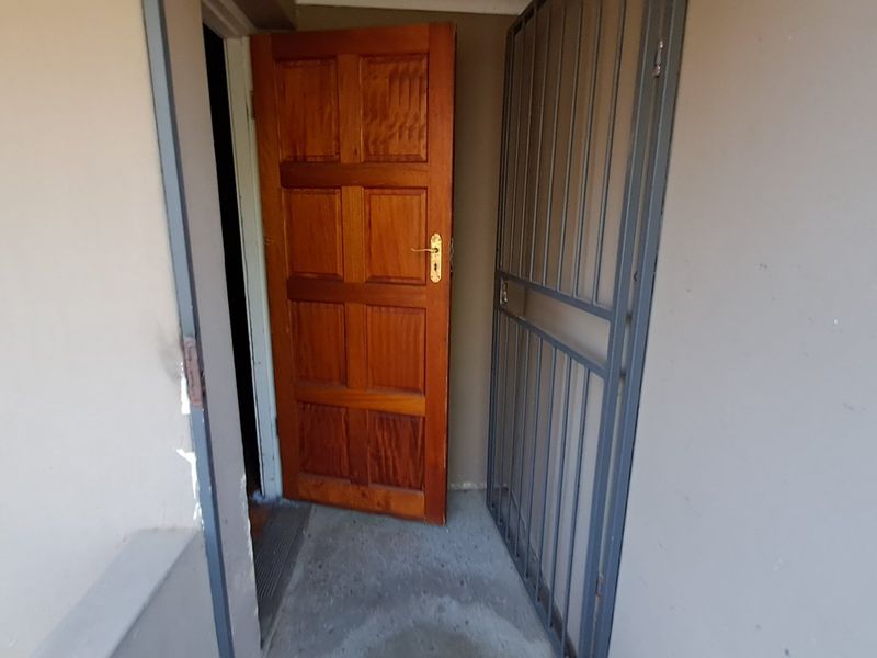 An exterior entrance area featuring a solid timber door and a metal security gate, showing signs of wear on the surrounding wall and floor.