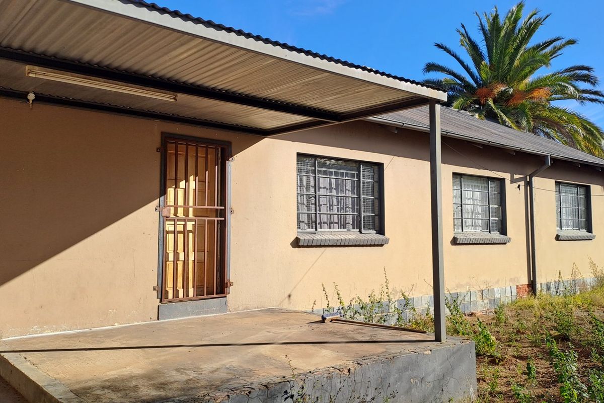 Rear exterior view of the house featuring a covered concrete patio, security gates, and window bars, with a weathered facade and surrounding garden area.