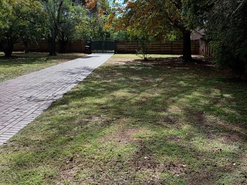 A spacious front garden featuring a paved driveway, mature trees, and secure perimeter fencing with a metal entrance gate.