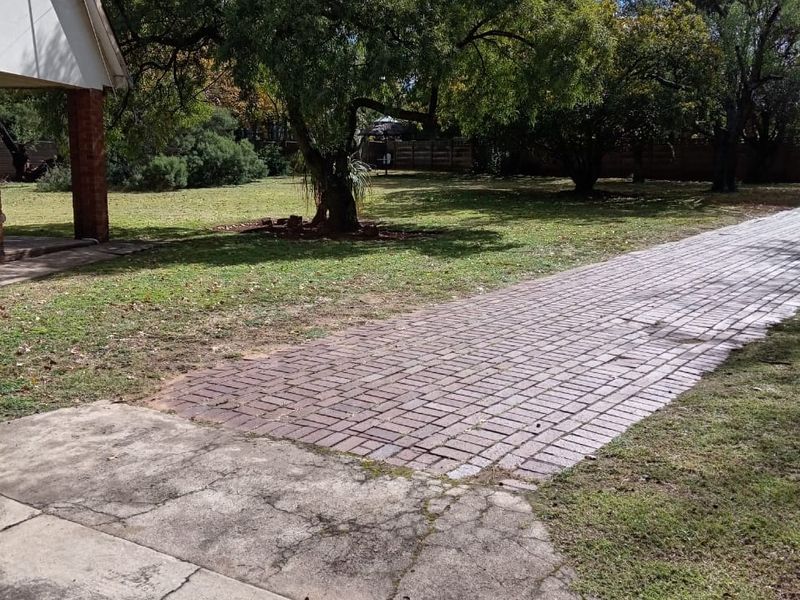 A spacious rear garden area featuring a brick-paved driveway, a concrete patio section, and established mature trees.