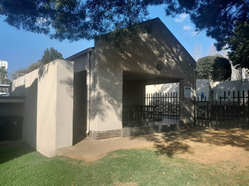 A covered outdoor communal area featuring tiled flooring and metal security fencing, situated within a landscaped garden setting.
