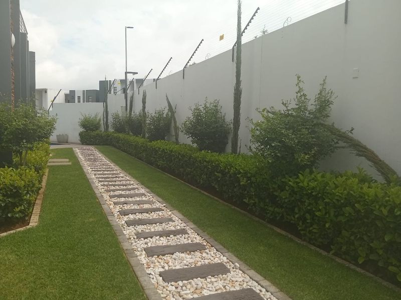 A well-maintained garden area featuring a stone and paver pathway, manicured hedging, and a secure perimeter wall with electric fencing.