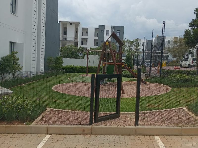 A well-maintained communal garden area featuring a fenced playground, adjacent to apartment building parking facilities.