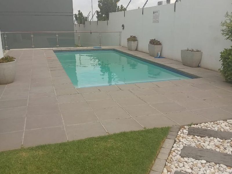 A well-maintained outdoor pool area featuring a tiled surround, glass balustrade, and landscaped garden elements with secure boundary walling.