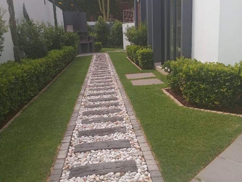 A well-maintained rear garden featuring a stone pathway, manicured lawn, perimeter hedging, and a built-in outdoor braai area.