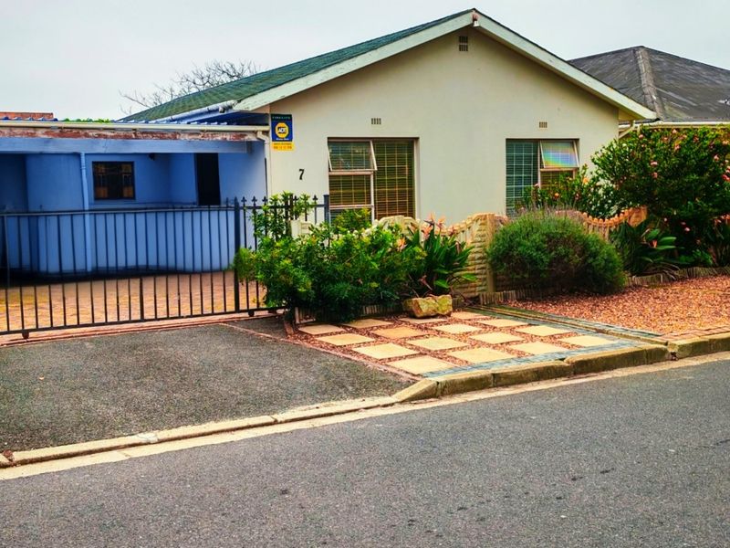 Front elevation of a single-story house featuring a paved driveway, metal security gate, and carport area with established garden landscaping.