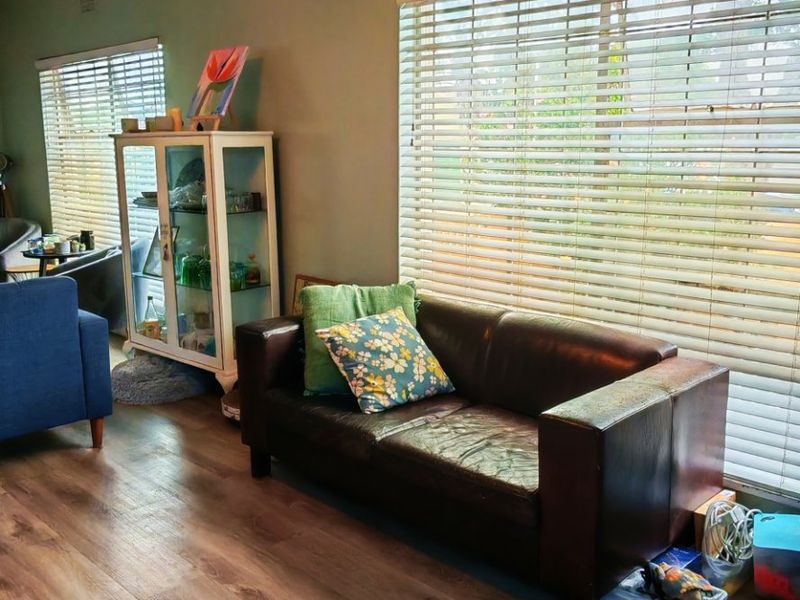A living room featuring laminate flooring, ceiling fan, and large windows with fitted venetian blinds.