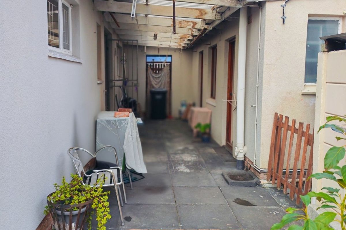A narrow rear exterior walkway featuring paved flooring and a weathered, partially covered roof structure requiring maintenance.