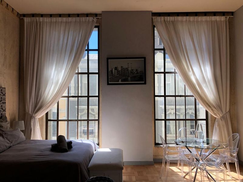 A well-lit bedroom featuring classic parquet flooring and large, multi-pane windows that provide ample natural light and urban views.