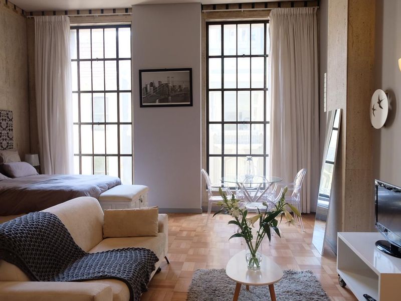 A bright open-plan living and bedroom area featuring parquet flooring, large industrial-style steel-framed windows, and exposed concrete structural columns.
