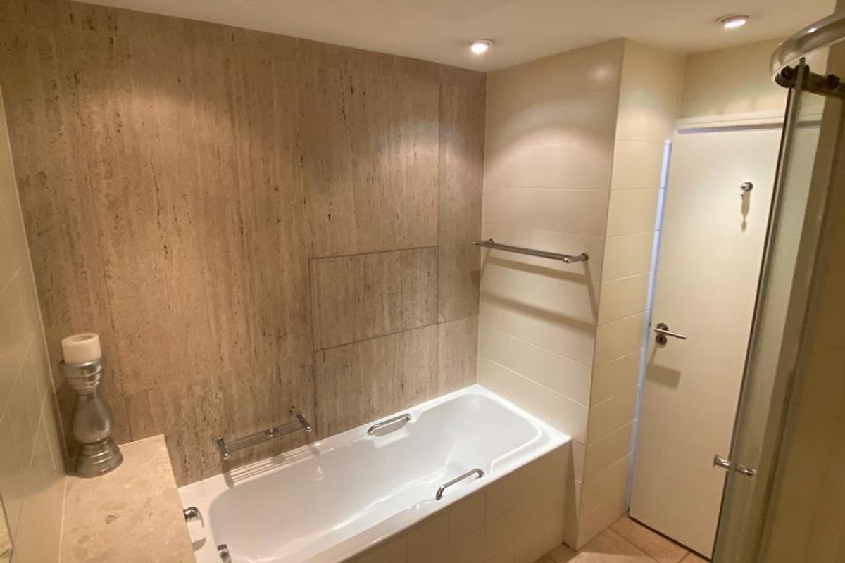 A well-maintained bathroom featuring a built-in bathtub, separate shower enclosure, pedestal washbasin, and neutral stone-look wall and floor tiling.
