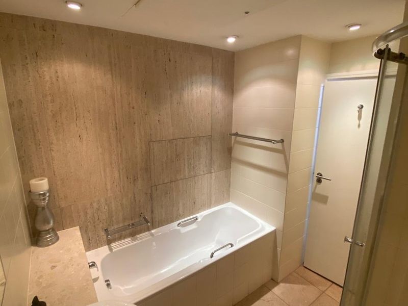 A well-maintained bathroom featuring a built-in bathtub, separate shower enclosure, pedestal washbasin, and neutral stone-look wall and floor tiling.