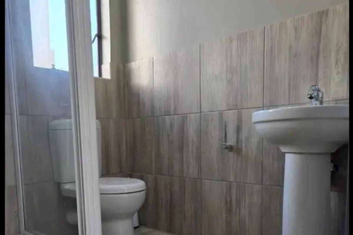 A clean, modern bathroom featuring a pedestal basin, toilet, and a glass-enclosed shower with neutral wood-look wall and floor tiles.