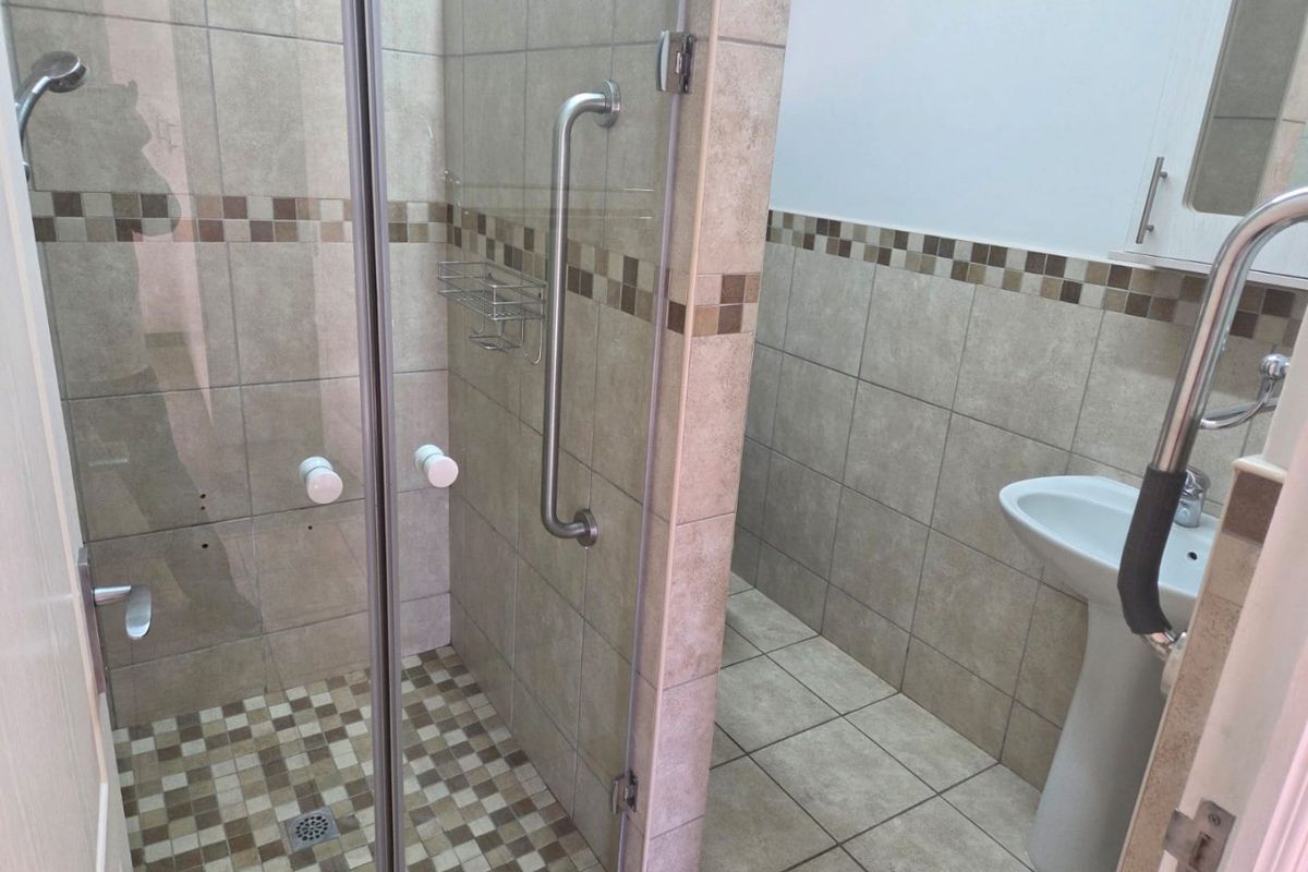 A functional bathroom featuring a glass-enclosed shower, pedestal basin, and safety grab rails with neutral ceramic tiling throughout.