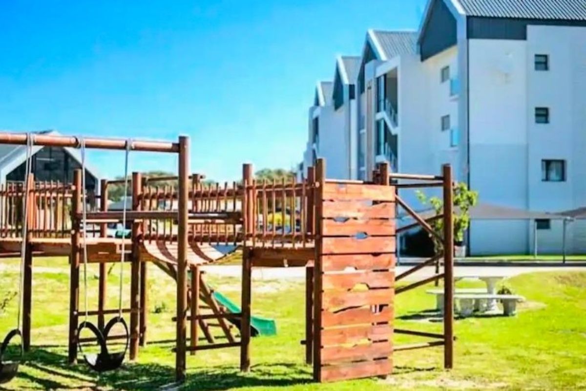 Communal outdoor area featuring a wooden playground structure set within a landscaped lawn, adjacent to the apartment building complex.