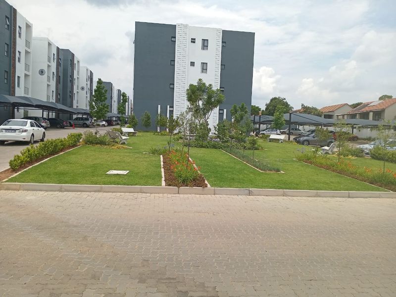 A well-maintained communal garden area within an apartment complex, featuring landscaped lawns, paved pathways, and covered parking bays.