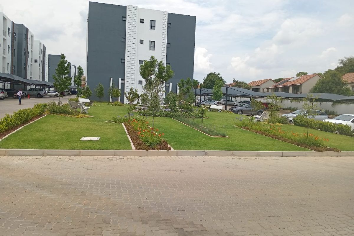 A well-maintained communal garden area featuring landscaped lawns and flower beds, situated between modern apartment blocks with covered parking facilities.