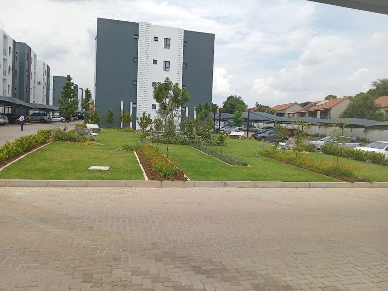 A well-maintained communal garden area featuring landscaped lawns and flower beds, situated between modern apartment blocks with covered parking facilities.