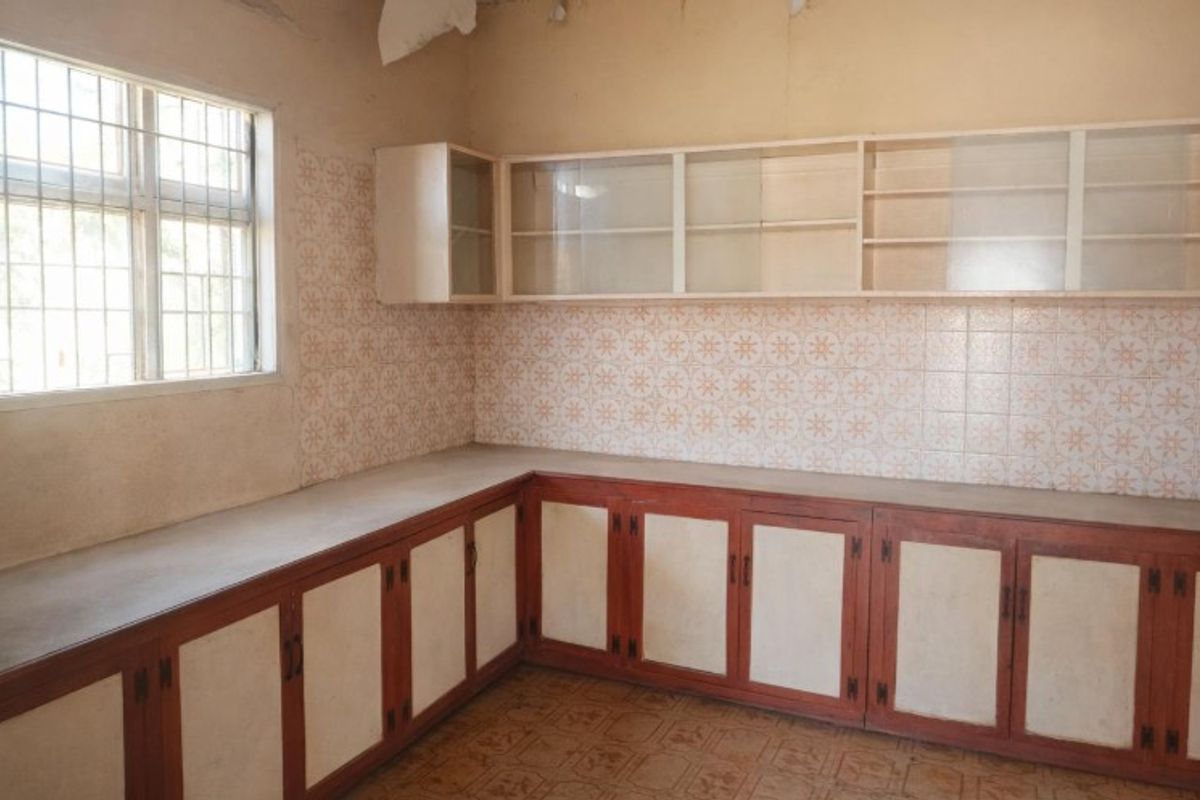 A dated, empty kitchen featuring original built-in cabinetry, tiled backsplash, and a window with security bars, requiring renovation to modernize the space.