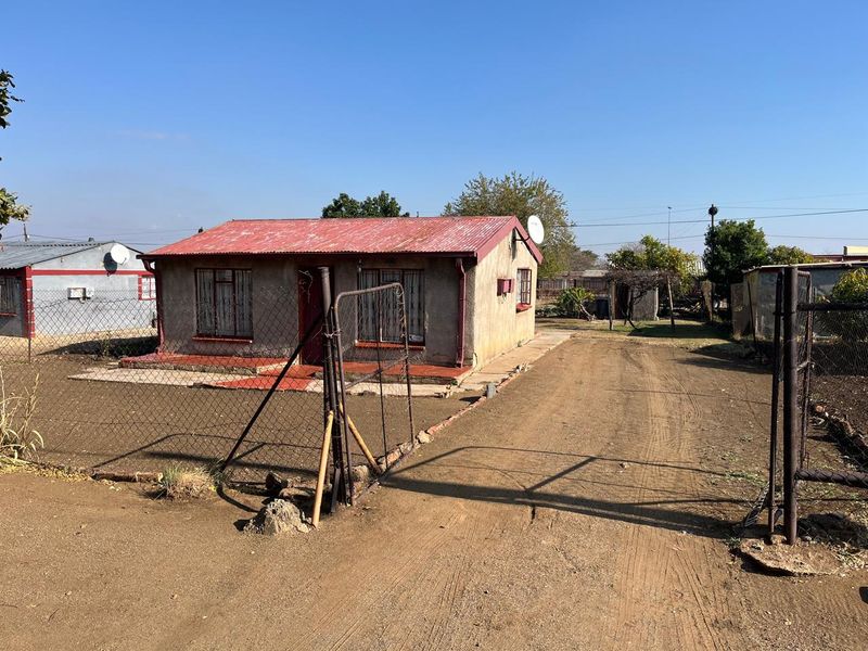 Front view of a detached house featuring a corrugated metal roof, security bars on windows, and a fenced yard with an unpaved driveway.