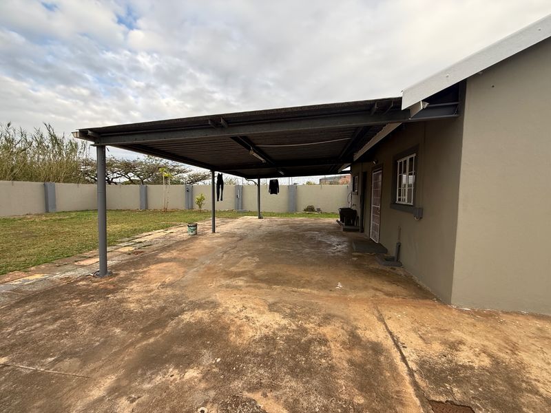 A spacious covered patio area featuring a steel roof structure and concrete flooring, overlooking a private garden with perimeter walling.