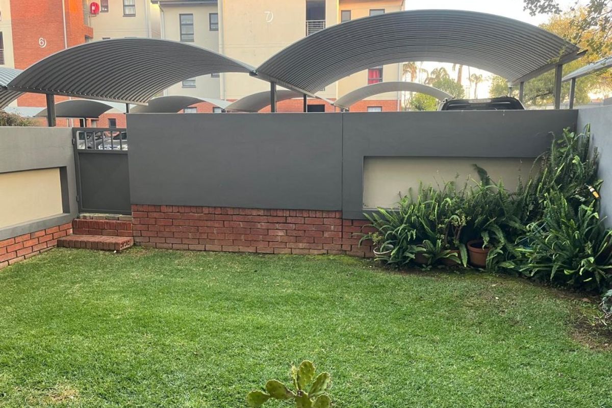 A private rear garden area featuring a well-maintained lawn, brick and plastered boundary walls, and a modern curved metal carport structure.
