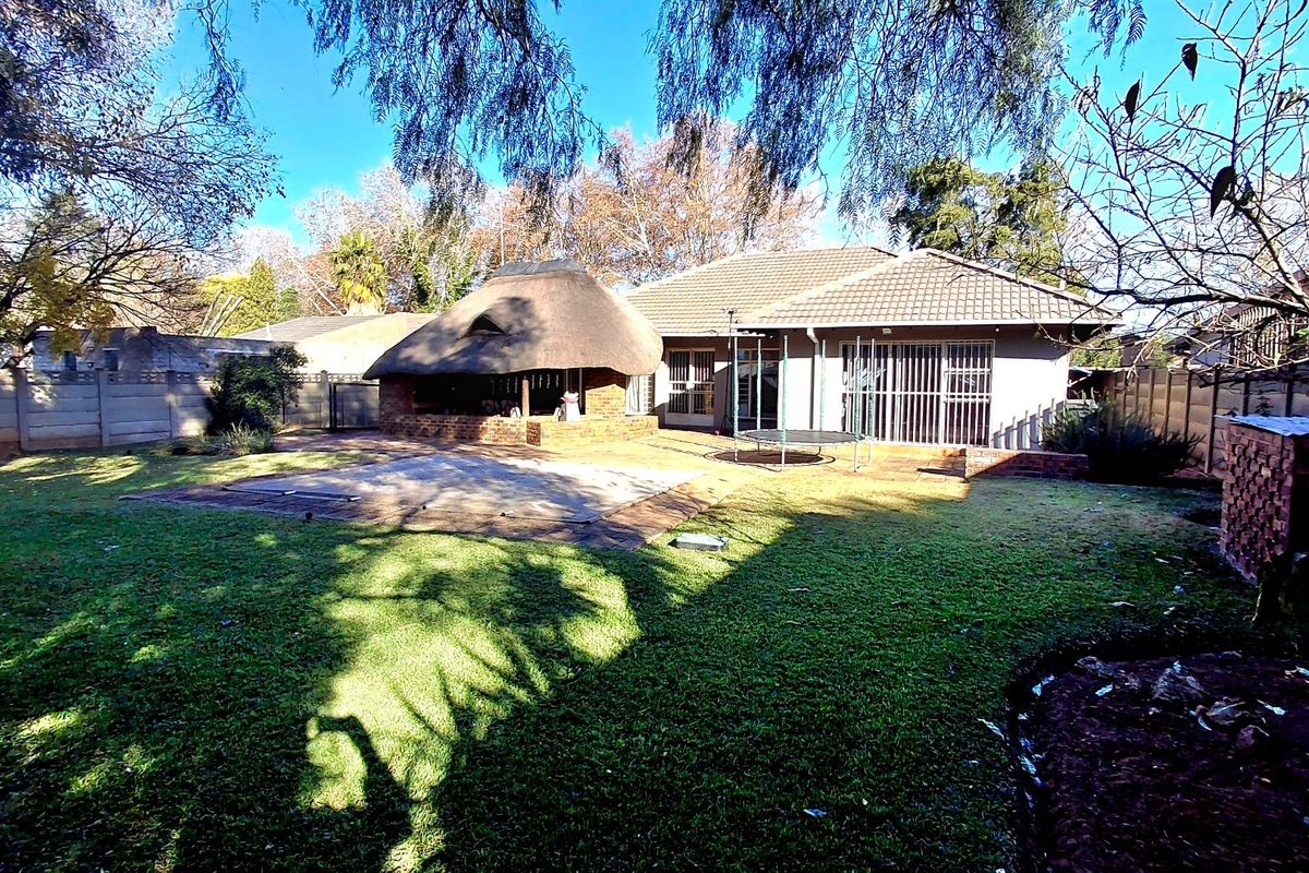 Rear garden view featuring a thatched lapa, swimming pool with cover, and a tiled roof house with sliding glass doors and security gates.