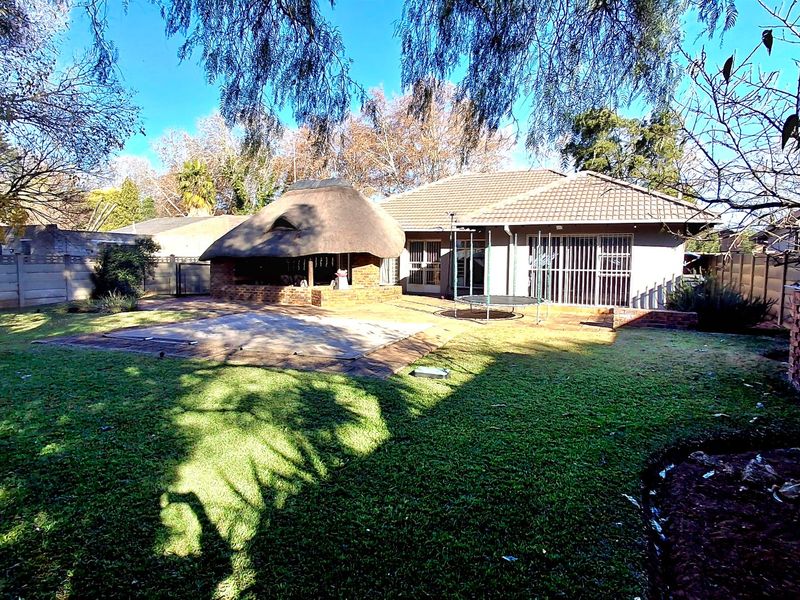 Rear garden view featuring a thatched lapa, swimming pool with cover, and a tiled roof house with sliding glass doors and security gates.