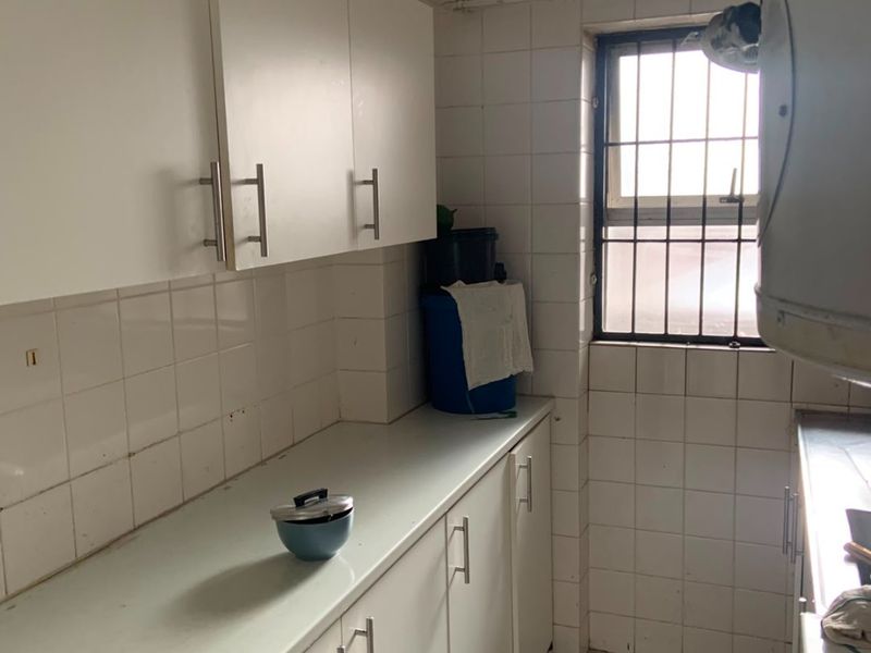 A compact kitchen featuring white built-in cabinetry, tiled walls and flooring, and a window with security bars.