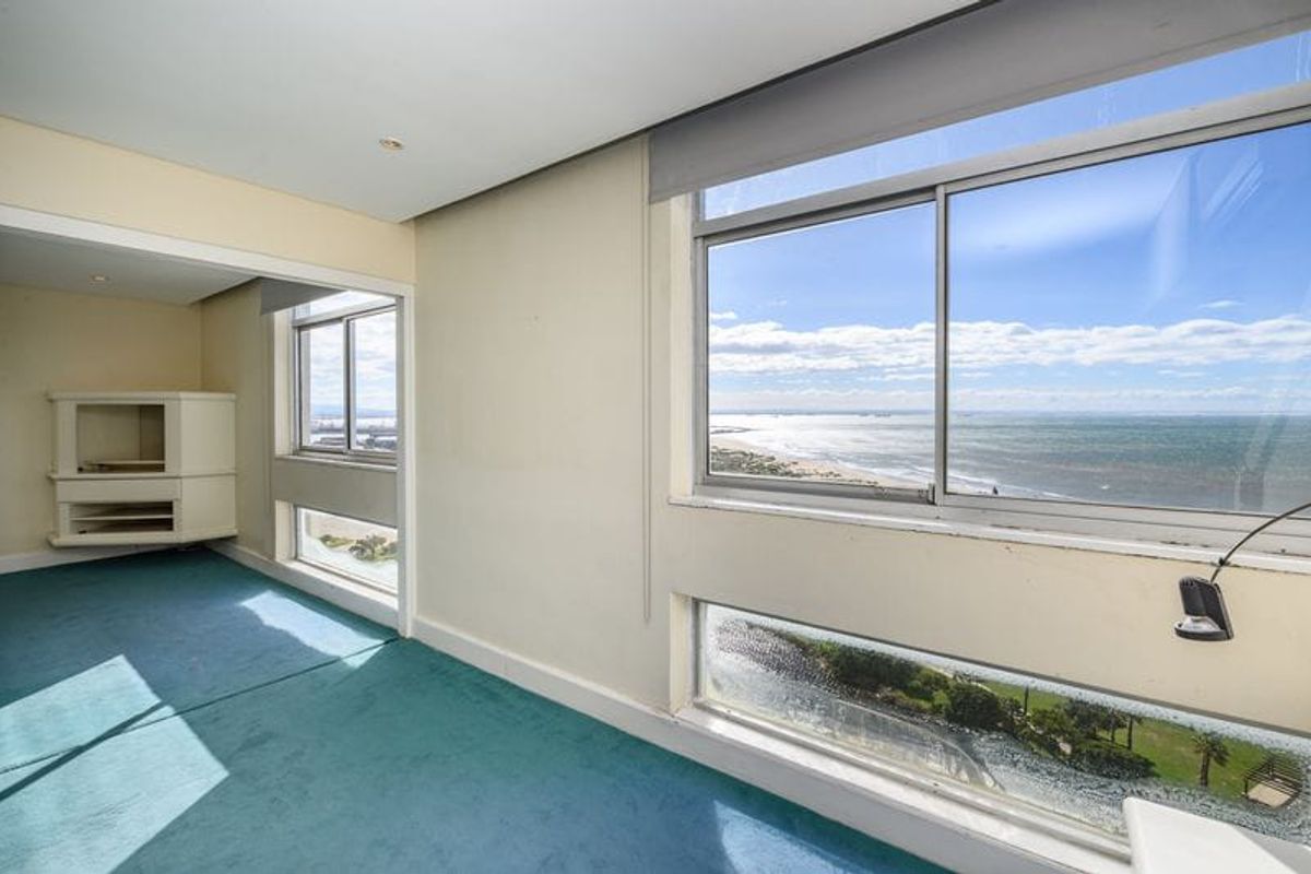 An empty living area featuring expansive windows with panoramic ocean views, built-in corner shelving, and recessed lighting, offering significant potential for modernization.