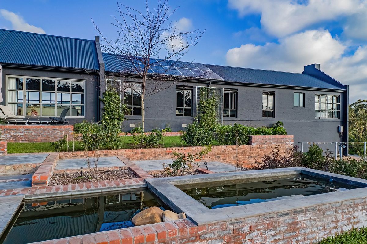 A well-maintained rear exterior view featuring a dark-painted farmhouse with solar panels, tiered brick garden beds, and a modern water feature.