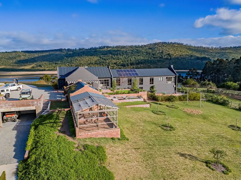 Aerial view of a modern farmhouse featuring a metal roof with solar panels, brick retaining walls, a garage, and expansive landscaped grounds.