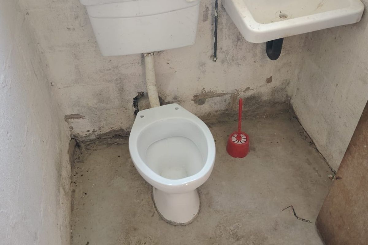 A basic, unfinished bathroom featuring a standard toilet and wall-mounted basin with exposed plumbing and raw concrete flooring.