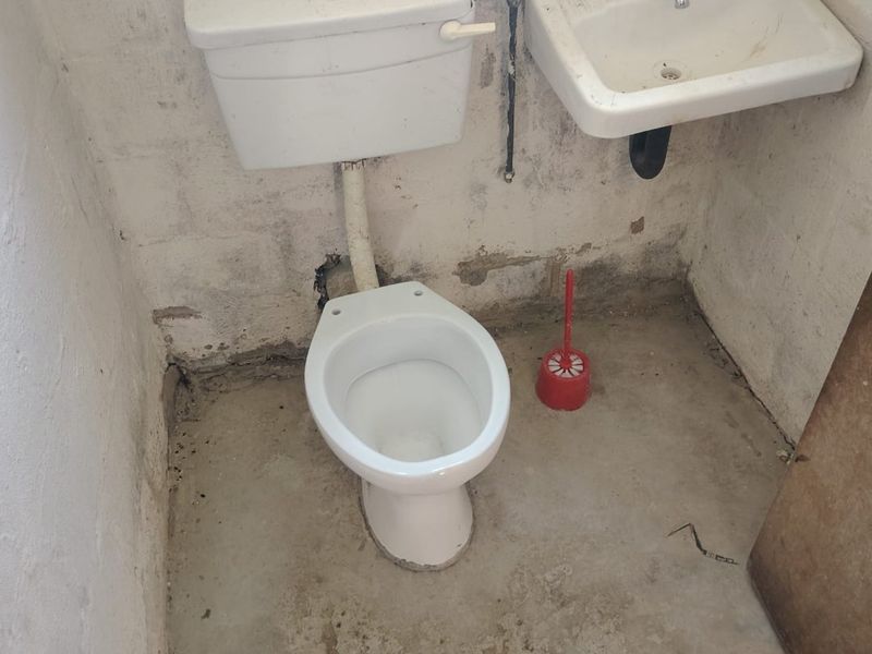 A basic, unfinished bathroom featuring a standard toilet and wall-mounted basin with exposed plumbing and raw concrete flooring.