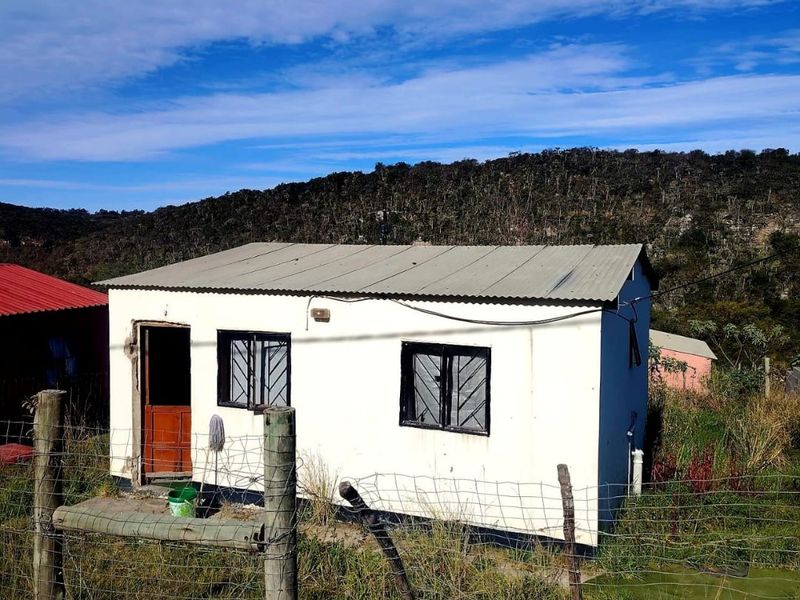 A detached single-story dwelling featuring a corrugated metal roof and security-barred windows, set within a fenced plot in a rural landscape.
