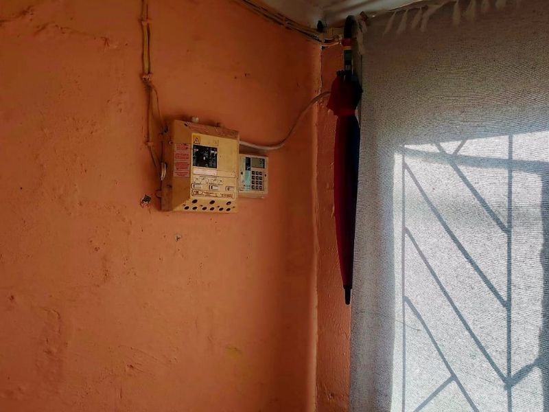 A close-up view of an interior wall featuring an exposed electrical distribution board, security alarm keypad, and window with security burglar bars.