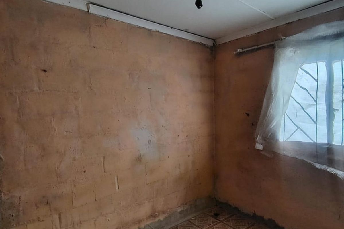 An empty bedroom featuring exposed brick walls, tiled flooring, and security bars on the window, requiring significant renovation and finishing work.