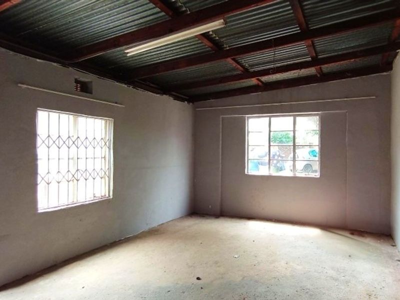 An empty, unfinished living area featuring exposed timber rafters, corrugated metal ceiling, and security burglar bars on the windows.