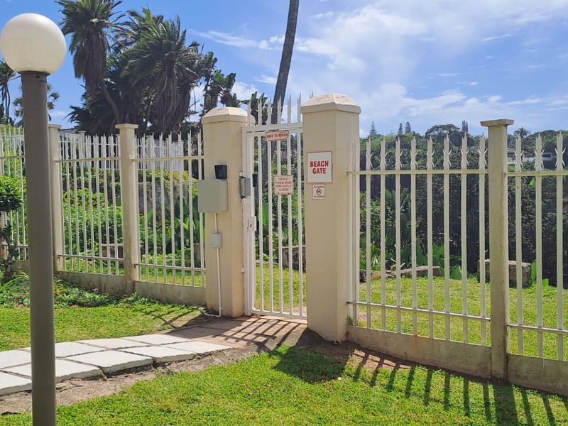 A secure pedestrian beach gate with metal fencing, concrete pillars, and a paved walkway leading through the landscaped grounds of the apartment complex.