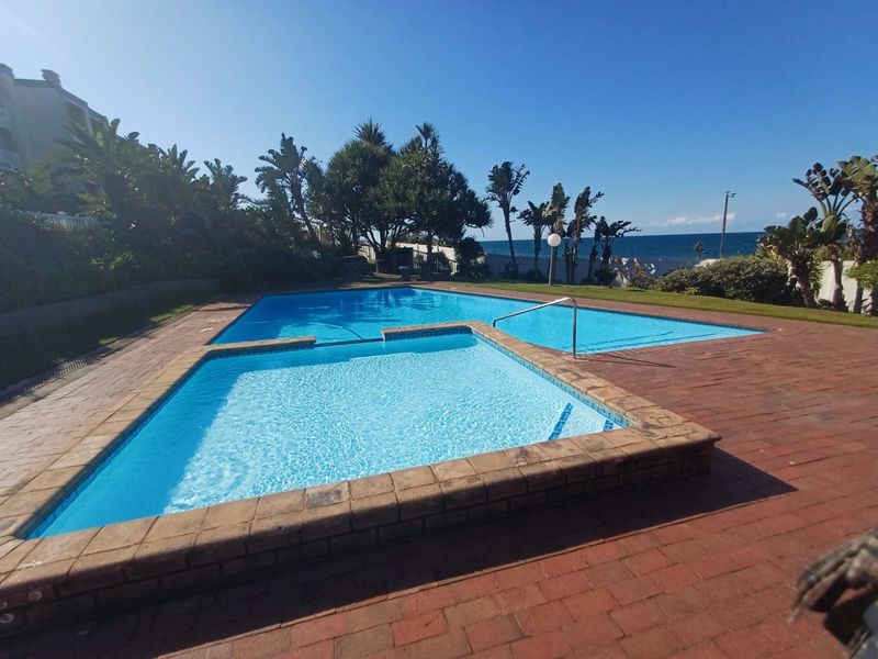 A well-maintained communal swimming pool area featuring paved decking, landscaped surroundings, and direct views towards the nearby coastline.