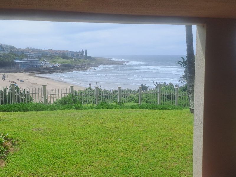 A scenic view of the coastline and beach from a private patio area with a well-maintained lawn and perimeter fencing.