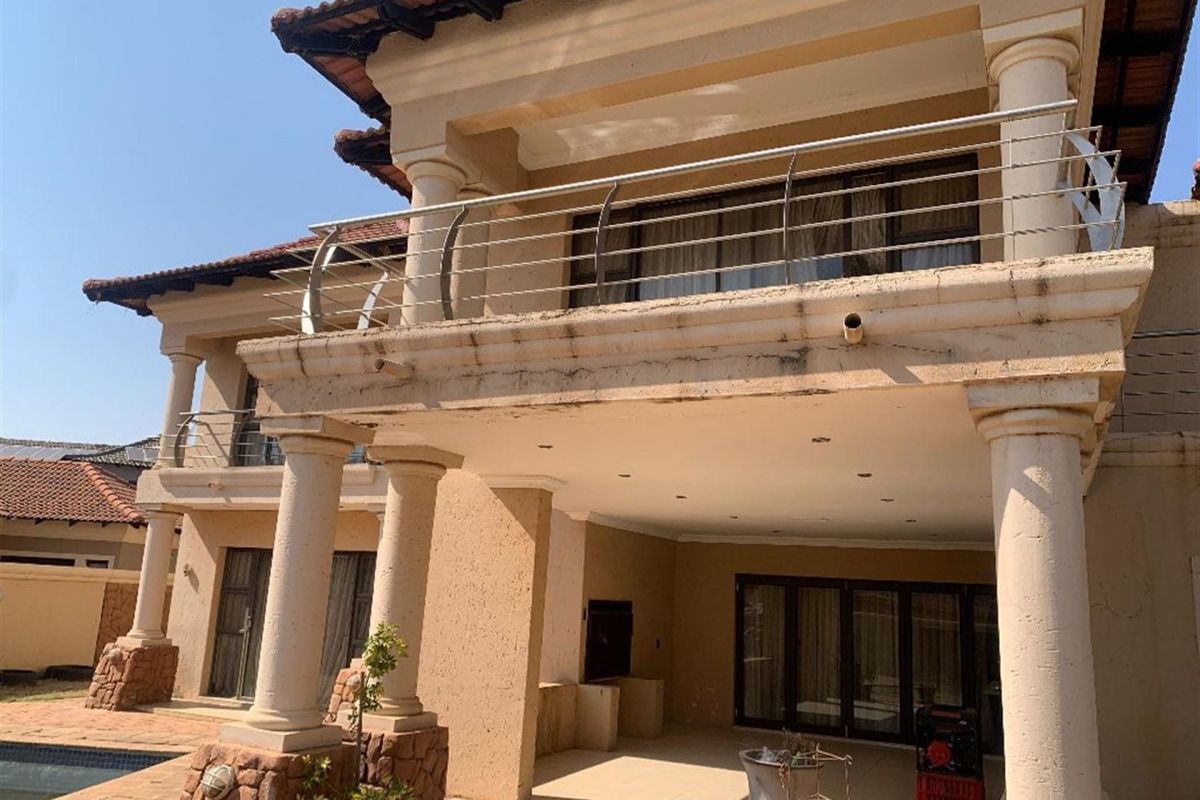 Rear elevation featuring a covered patio with built-in braai, upper balcony with stainless steel balustrade, and classic architectural columns.