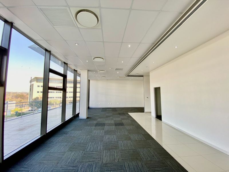 A bright, vacant commercial office space featuring floor-to-ceiling windows, suspended ceilings with recessed lighting, and a mix of carpet and ceramic tile flooring.