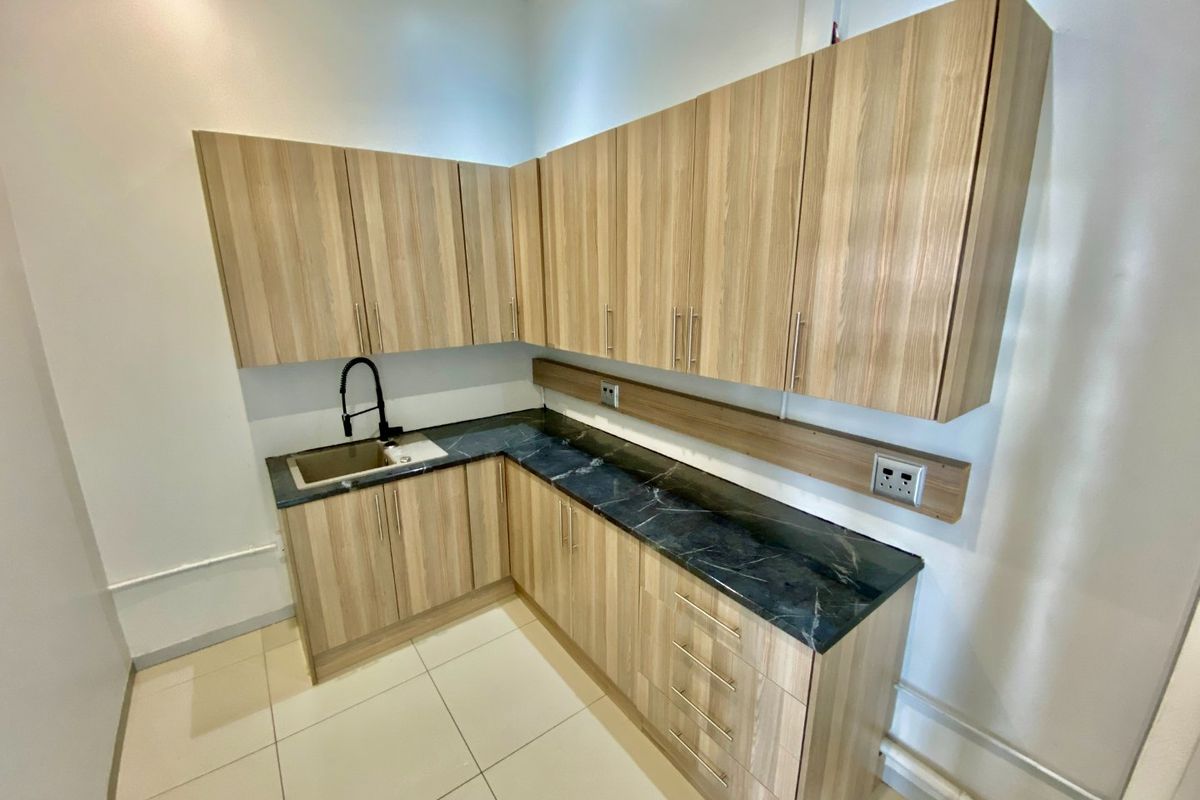 A modern, clean kitchenette featuring light wood-grain cabinetry, dark stone countertops, and a sleek black gooseneck faucet in a bright, tiled space.