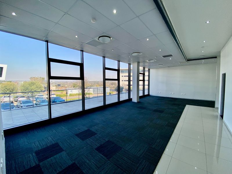 Spacious, modern open-plan office area featuring floor-to-ceiling windows, recessed lighting, and a combination of carpet and ceramic tile flooring with balcony access.