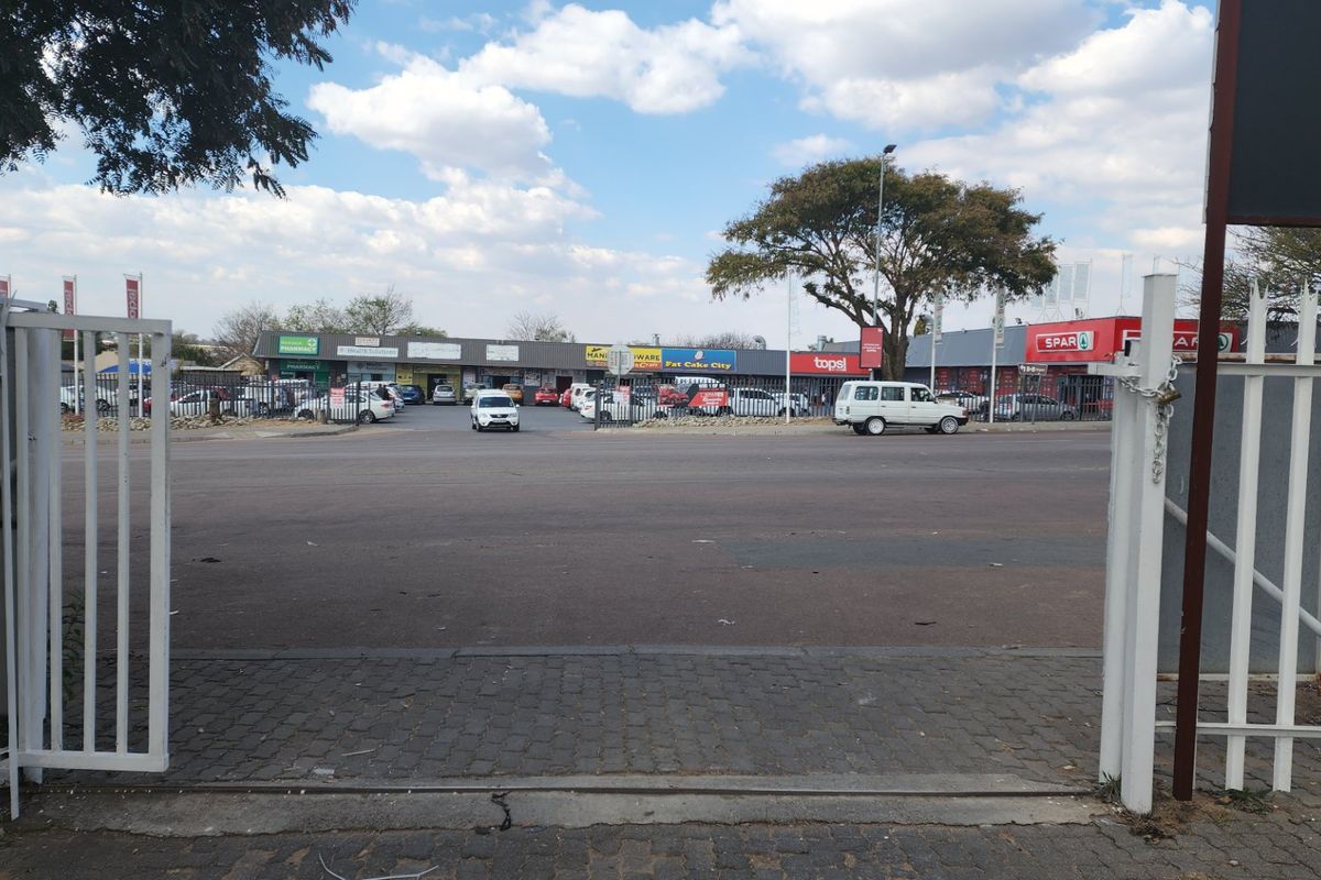 View from a gated entrance looking across a street towards a commercial shopping center with various retail storefronts.