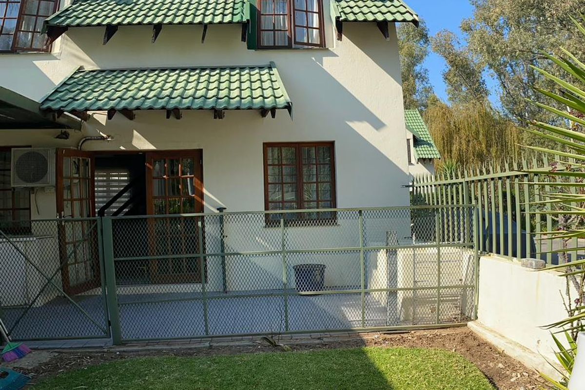 Rear view of a townhouse featuring a tiled roof, timber frame windows and doors, a paved patio area, and a private lawn with security fencing.