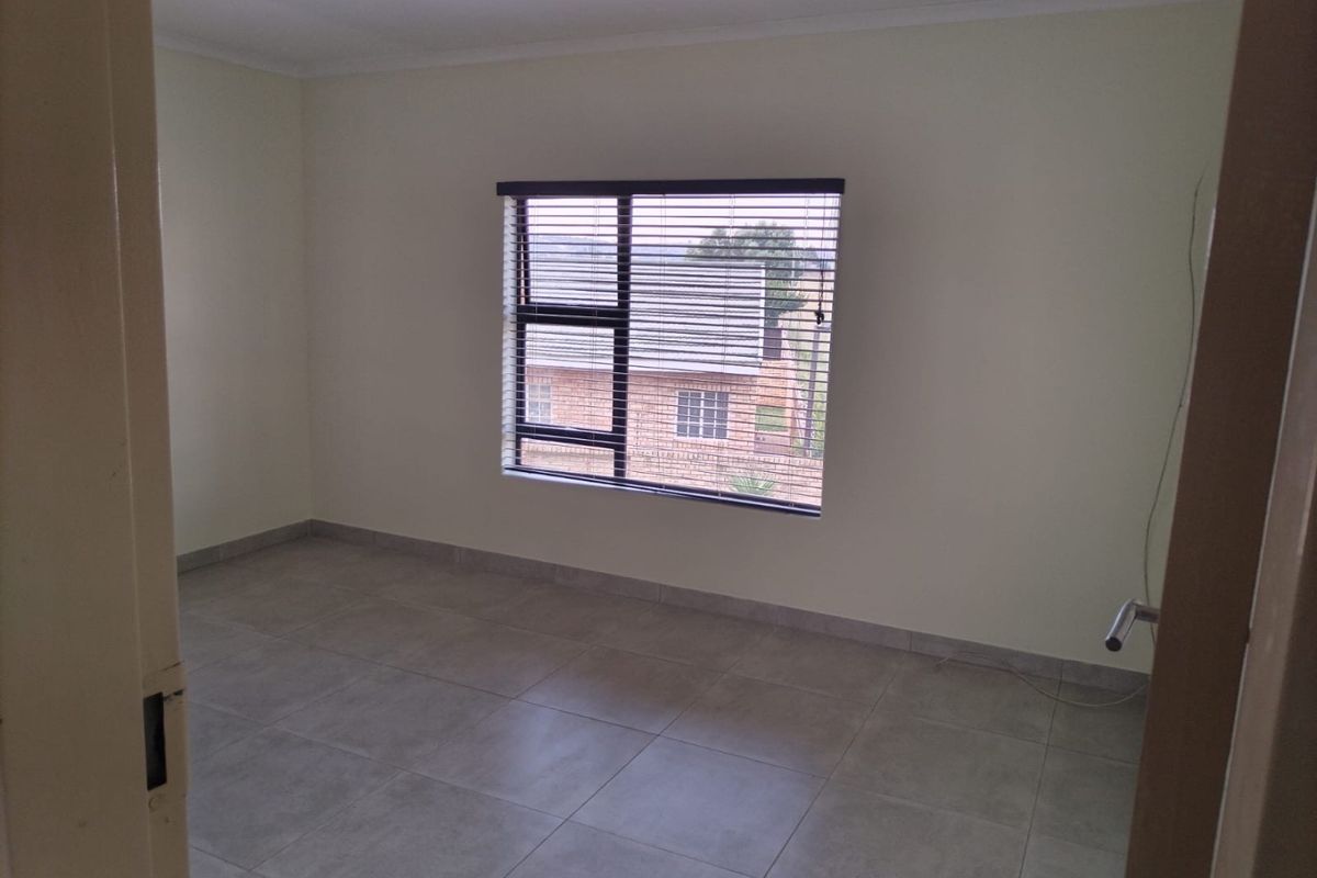 A clean, empty bedroom featuring neutral tiled flooring and a large window with fitted venetian blinds, offering a bright and versatile living space.