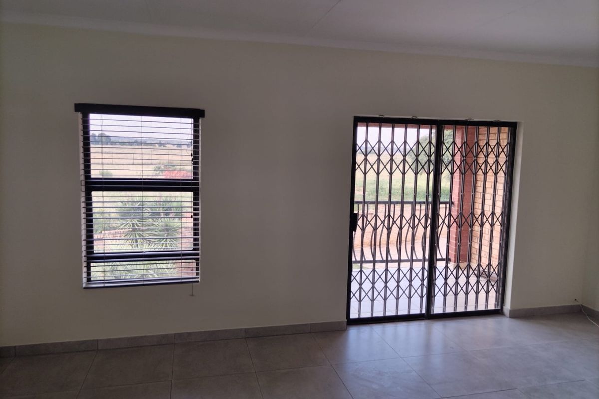 A bright, empty living area featuring neutral tiled flooring, a sliding glass door with a security gate, and a window with fitted blinds.