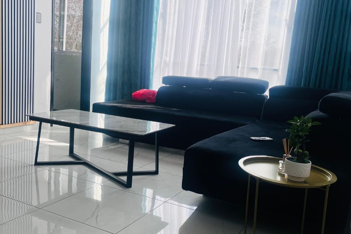 A bright living room featuring polished marble-look floor tiles, decorative wall paneling, and large windows with floor-to-ceiling curtains.
